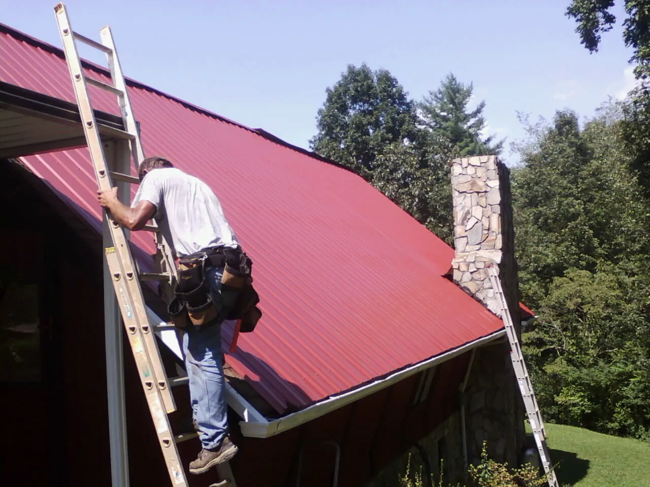 Roofer on a ladder beside a red metal roof and stone chimney — Soffit & Fascia Repair by Roofing Brothers in Kathryn